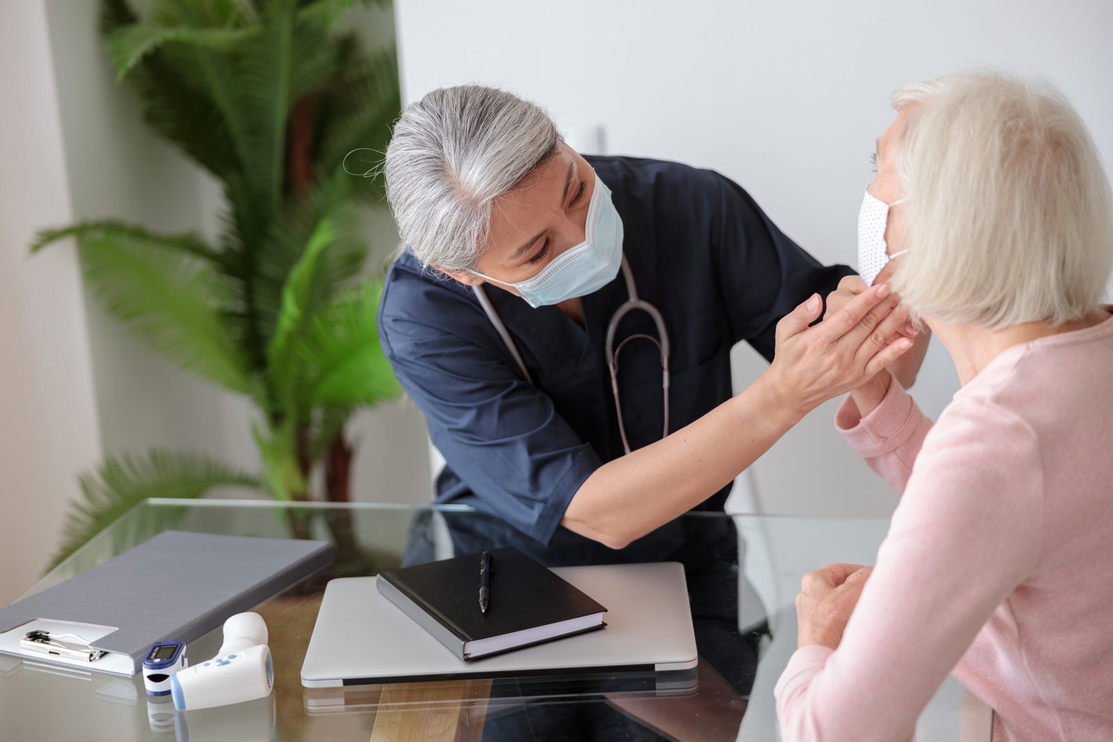 Medical practitioner looking at patient and checking lymph nodes in the neck, elderly lady sitting on sofa in protective masks