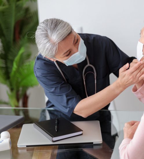 Medical practitioner looking at patient and checking lymph nodes in the neck, elderly lady sitting on sofa in protective masks
